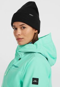 Giacca impermeabile color turchese con colletto alto, chiusura con zip e logo. Cappello beanie a coste neri. Texture liscia e colori vivaci.