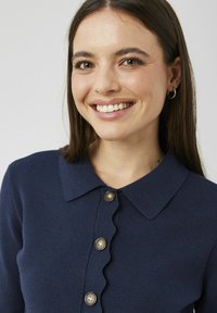 Jeune femme aux longs cheveux bruns et aux boucles d'oreilles créoles souriante, portant un pull bleu marine avec une patte de boutonnage à bords festonnés et un col.