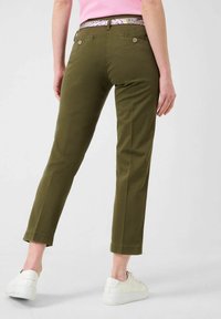 Olivgrüne, taillierte Hose mit glatter Textur, ausgestattet mit zwei Gesäßtaschen und einem bedruckten Bund, kombiniert mit weißen Sneakers.