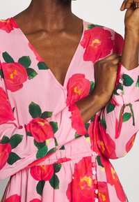 Rosa Blumenkleid mit großen roten Rosen und grünen Blättern, mit tiefem V-Ausschnitt, langen Ärmeln und Knopfakzenten an der Manschette.