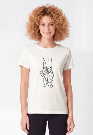 Femme aux cheveux bouclés portant un t-shirt blanc avec un dessin au trait noir de pointes de ballet croisées sur le devant.