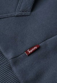 Mørkeblå strikket stof med et rødt rektangulært mærke mærket "Superdry", syet nær en lomme søm.