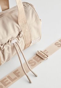 Vaaleanbeige nylonista valmistettu duffel-laukku, jossa on nyörikiinnitys, kudottu olkahihna, jossa lukee "SELF", sekä muovinen solki. Sileä, matta pinta.