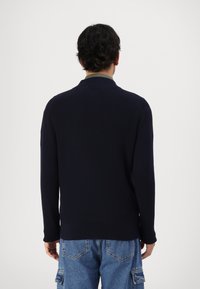 Pulover tricotat bleumarin cu o textură ușor cu riburi, având un guler fals și mâneci lungi, asortat cu pantaloni cargo de culoare bleumarin deschis.