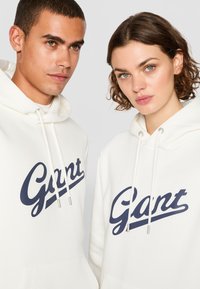 Hvite hettegensere med en blå "Gant" logo over brystet, med snor og frontlommer, brukt av to personer side om side.