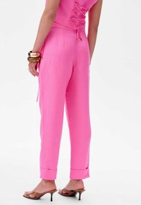 Paarse jumpsuit met een gekruist rugdetail, taps toelopende broekspijpen en opgerolde manchetten. Geaccessoreerd met gestapelde armbanden en bruine haksandalen.