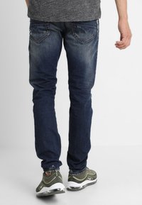 Jeans de mezclilla de un azul oscuro con un corte ajustado, que presentan un sutil desgaste y efectos de arrugas. Los bolsillos traseros tienen acentos de costura distintivos.