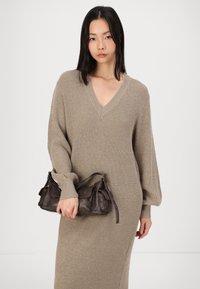 Beige ribbad stickad lång klänning med V-ringning, med långa puffy ärmar. Modellen håller en mörkbrun läderhandväska med en strukturerad design.