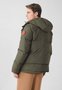 Chaqueta acolchada verde oliva con capucha, que presenta un parche redondo rojo en el hombro y una textura acolchada, llevada sobre pantalones beige.