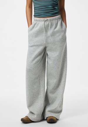Personne portant un pantalon de jogging ample gris clair avec cordon de serrage, un top court rayé bleu et noir, et des chaussures marron, debout les mains dans les poches.
