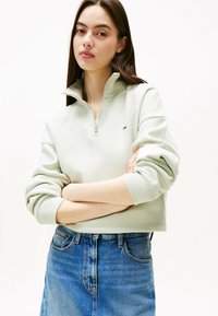 Felpa verde chiaro con zip, colletto alto e polsini a coste, abbinata a una gonna di jeans blu con una leggera forma ad A.