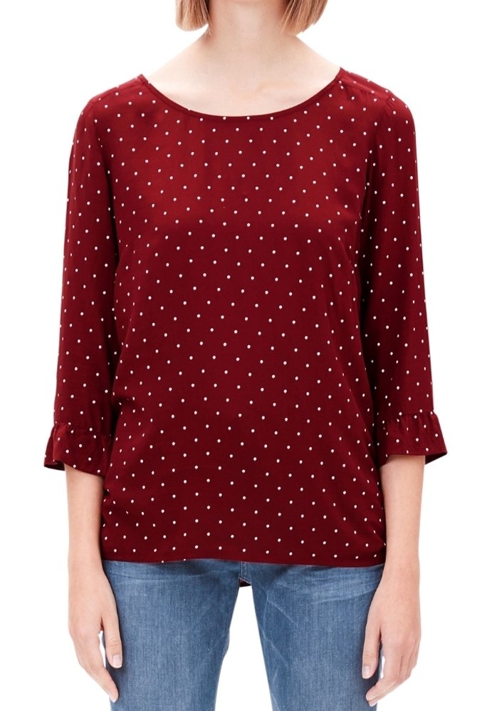 Femme portant un chemisier bordeaux à manches longues avec des pois blancs et un jean bleu, debout sur un fond blanc.
