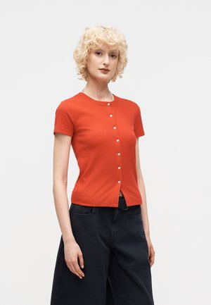 Rövid, göndör szőke hajú nő, aki szűk, rövid ujjú, piros, gombos felsőt és bő fekete nadrágot visel, sima háttér előtt áll.