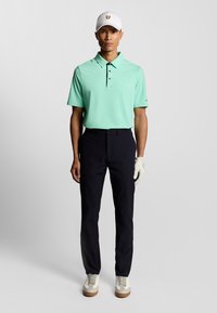 Ljusgrön polo med krage, svarta byxor och vita golfsandaler. Bär också en vit keps med en gul logotyp och en golfhandske.