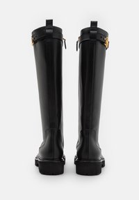 Bottes noires montant jusqu'au genou en cuir lisse avec une semelle épaisse, dotées d'une fermeture éclair latérale et d'une sangle décorative avec une boucle de couleur dorée.