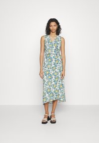 Robe portefeuille à motifs floraux avec un décolleté en V, motif de fleurs teal et jaunes, longueur midi, et une ceinture à nouer. Associée avec des sandales noires à lanières.