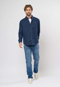 Tamsiai mėlynas ilgomis rankovėmis marškinis su klasikiniu apykakle, dėvimas virš baltos marškinėlių, derinamas su šviesiai mėlynomis džinsinėmis kelnėmis ir neutralių spalvų sportbačiais.