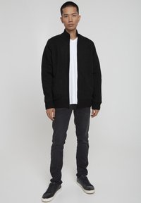 Cardigan negru din tricot cu guler înalt și manșete cu dungi, purtat peste un tricou alb cu decolteu în V, asortat cu jeansi închiși la culoare, slim fit, și adidași negri.