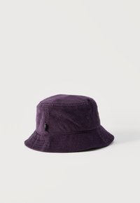 Σκούφο κουβαδάκι από κοτλέ σε σκούρο μωβ χρώμα με μια μικρή μαύρη ετικέτα στη μία πλευρά, τοποθετημένος επάνω σε ένα απλό ανοιχτόχρωμο φόντο.