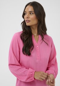 Femme aux longs cheveux noirs portant un chemisier rose vif à manches longues avec des boutons, ajustant sa manche, regardant à gauche sur un fond uni.