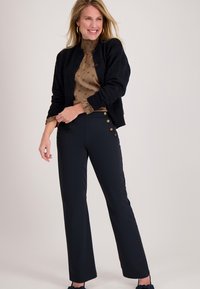 Zwarte cardigan, beige blouse met patroon en navy broek met gouden knoopaccenten. Gladde texturen en klassiek model; casual outfit.
