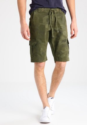 Homme portant un short cargo camouflage vert avec taille à cordon et des baskets blanches Nike, marchant sur fond blanc.