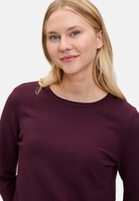 Pull à manches longues en bordeaux profond, fabriqué en tissu tricot doux. Il présente un col rond et une finition lisse et texturée.