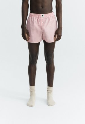 Homme portant un boxer rayé rose clair et blanc avec des chaussettes couleur crème, debout devant un fond blanc uni.