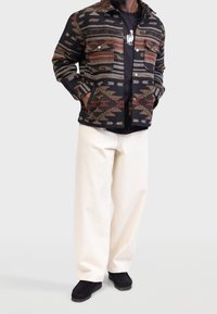 Giacca tessuta con un motivo geometrico multicolore nei toni del nero, marrone e grigio, abbinata a una maglietta nera e a pantaloni bianchi a gamba larga.