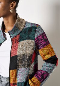 Osoba ubrana w wielokolorowy sweter z zamkiem błyskawicznym w stylu patchwork, biały podkoszulek, szare spodnie i złote kolczyki w kształcie kółek, trzymająca rękę w kieszeni.