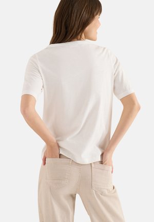 Femme aux cheveux bruns portant un t-shirt blanc uni à manches courtes et un pantalon beige, mains posées dans les poches arrière, tournée de dos.