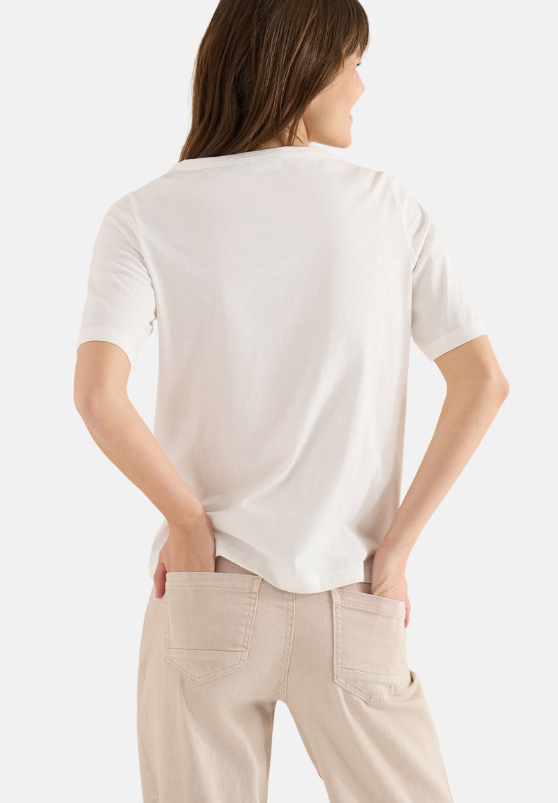 Femme aux cheveux bruns portant un t-shirt blanc uni à manches courtes et un pantalon beige, mains posées dans les poches arrière, tournée de dos.