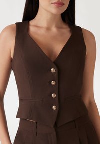 Bruine vest met een diepe V-halslijn, voorzien van vijf zilveren knopen, twee voorkruiszakken en een getailleerde pasvorm. Glad weefsel.