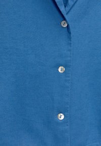 Camisa de lino azul con una textura suave, botonera con botones circulares y brillantes, y cuello doblado.