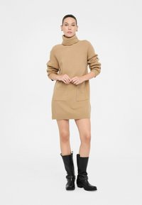 Abito in maglia beige con colletto alto a coste, maniche lunghe e tasche applicate sul davanti. Modello abbinato ad stivali neri al ginocchio.