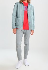 Chaqueta azul claro con botones, camiseta gráfica roja, jeans claros desgastados y zapatillas blancas. Atuendo casual para una figura masculina.