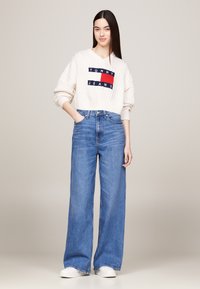 Weißer gerippter Pullover mit "TOMMY JEANS"-Logo, kombiniert mit hoch taillierten, weit geschnittenen blauen Jeans und weißen Sneakers.
