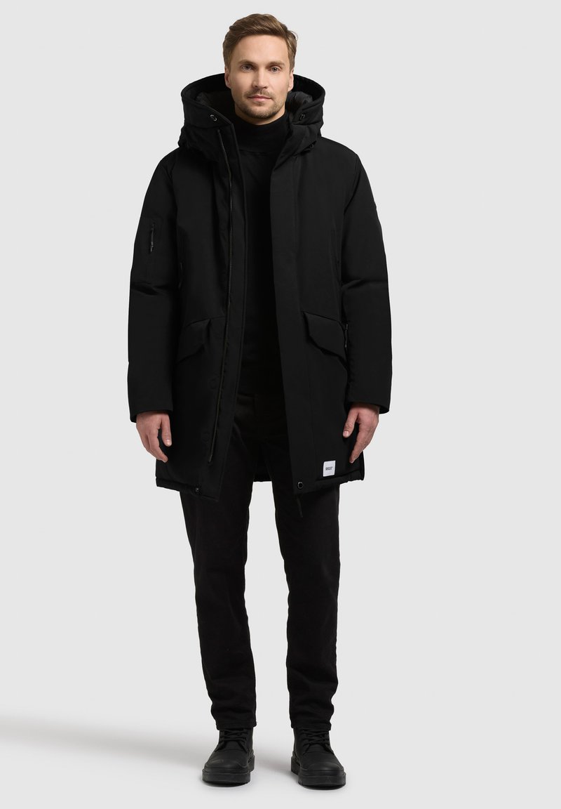 Sort parka med hætte, lynlåslukning, to forlommer og et mærkelogo ved kanten; båret over en sort turtleneck og bukser.