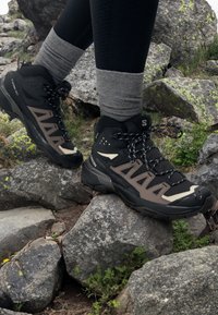 Des chaussures de randonnée noires et brunes avec un design montagnard, en matériaux synthétiques et en maille, et une semelle en caoutchouc robuste, posées sur un terrain rocheux.