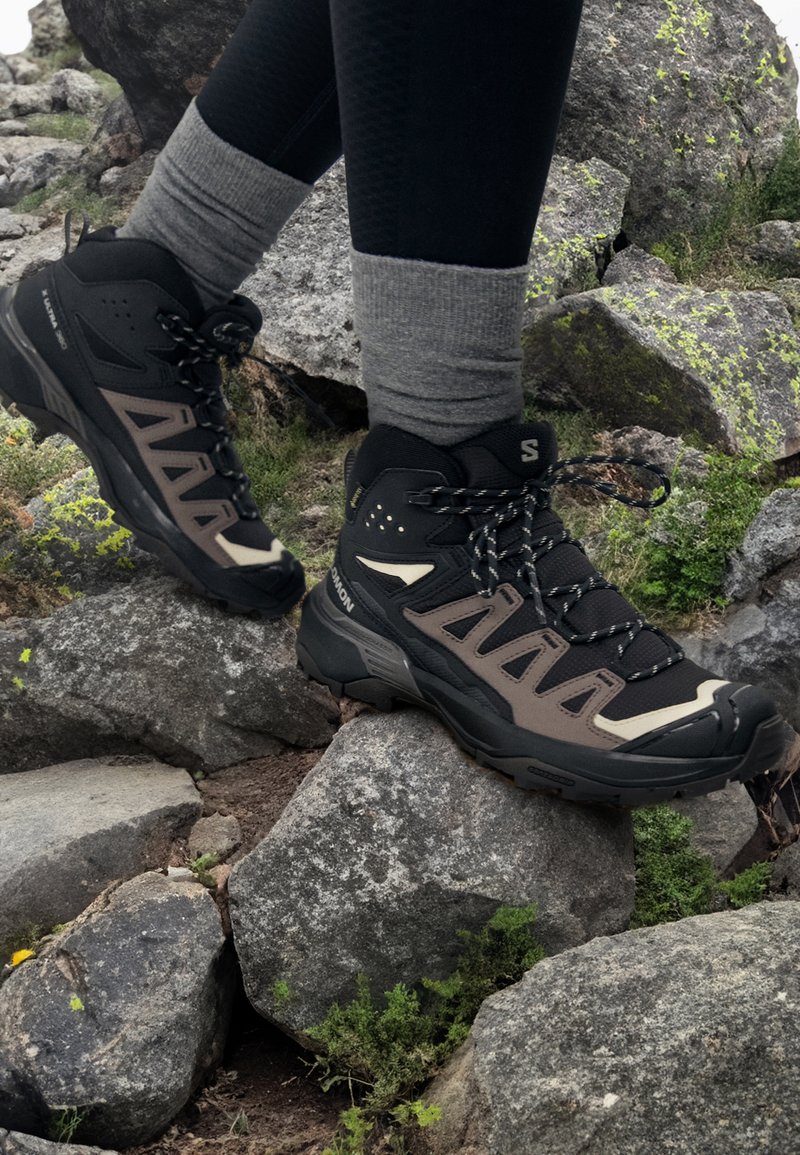 Des chaussures de randonnée noires et brunes avec un design montagnard, en matériaux synthétiques et en maille, et une semelle en caoutchouc robuste, posées sur un terrain rocheux.
