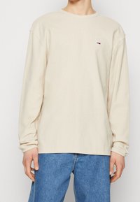 Beżowa koszula z długim rękawem, wykonana z miękkiej bawełny, z okrągłym dekoltem i prążkowaną strukturą. Posiada małe logo na lewej piersi. W zestawie z dżinsowymi spodniami.