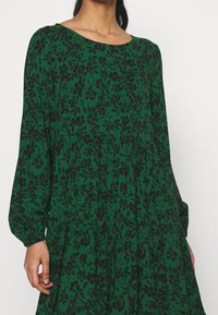 Vestido floral verde con patrones de flores negras, mangas largas y cuello redondo. Hecho de una tela ligera con una silueta fluida.