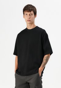 T-shirt nera oversize con maniche corte, colletto rotondo e tessuto morbido. Abbinata a pantaloni cargo grigi. Design semplice e minimal.