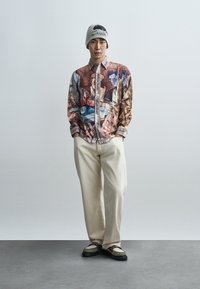 Graphic print shirt in multi-colors, beige baggy pants, and a gray beanie. Shoes feature a green and beige design with black soles.