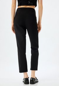 Pantalons noirs ajustés au design épuré, avec une texture lisse, une longueur courte et de subtils poches arrière, associés à des chaussures noires brillantes.