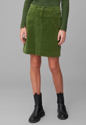 Jupe trapèze en velours côtelé vert avec poches devant et fermeture à boutons, portée avec des bottines noires et un haut vert côtelé à manches longues.