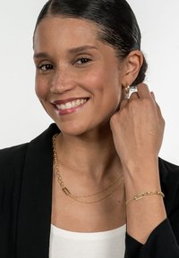 Gold layered chain necklace and bracelet with rectangular links. Model wears a black blazer and a white top, smiling at the camera.