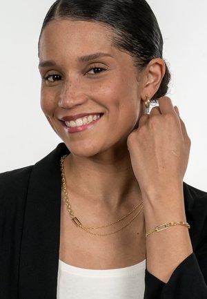 Gold layered chain necklace and bracelet with rectangular links. Model wears a black blazer and a white top, smiling at the camera.
