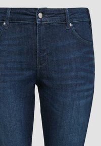 Ciemnoniebieskie dżinsy o gładkiej teksturze, z pięcioma kieszeniami i srebrnymi elementami na pasie. Posiadają klasyczny prosty krój.