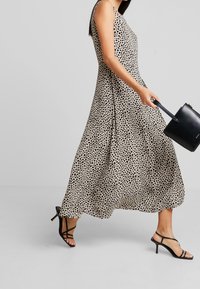 Beige sleeveless maxi dress dengan polka dot hitam, siluet yang mengalir, dipasangkan dengan sandal hitam bertali dan tas kecil hitam.